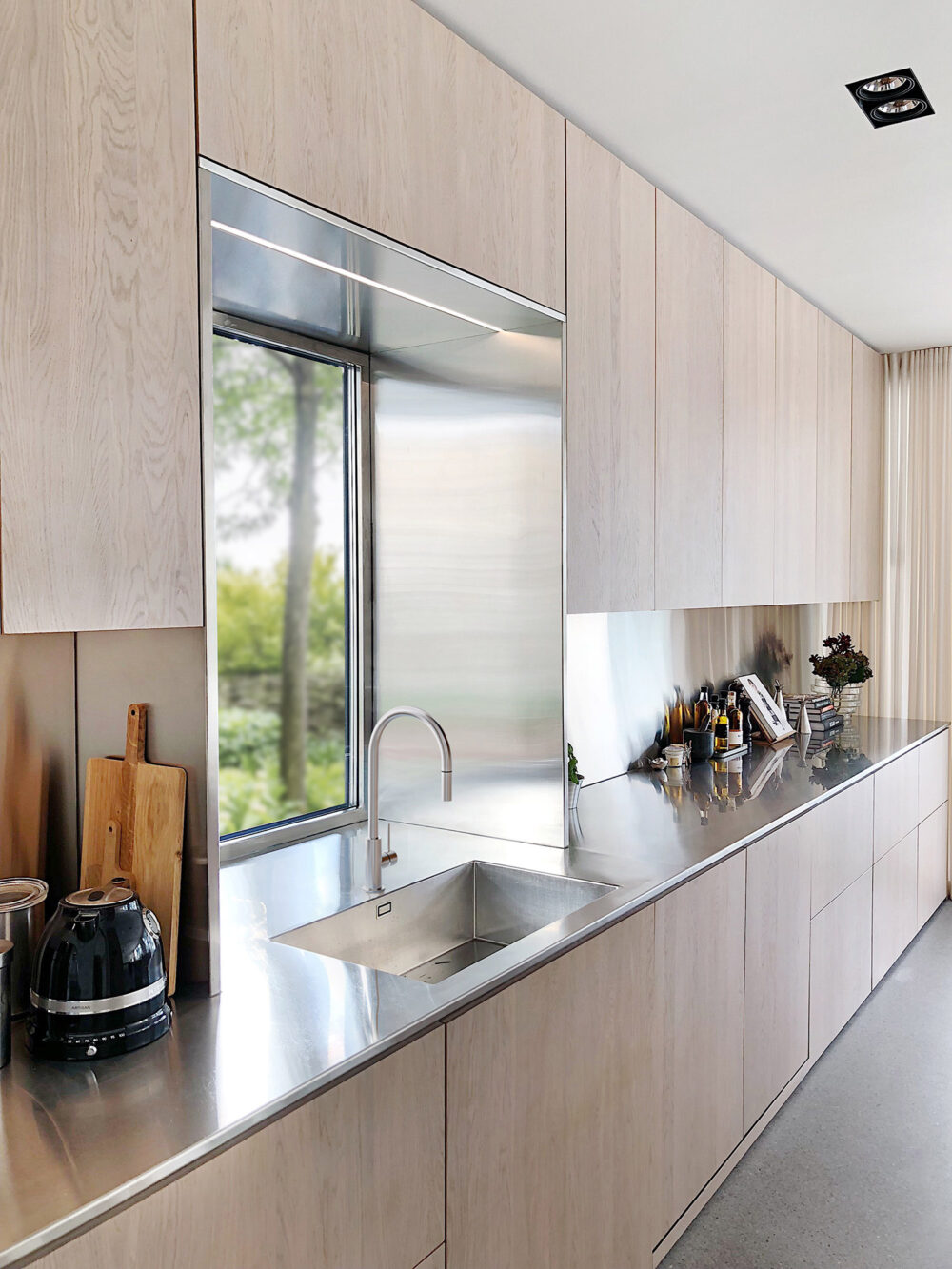 En del av ett kök med betonggolv, skåpsluckor i ljust trä och en rektangulär diskho i rostfritt stål.